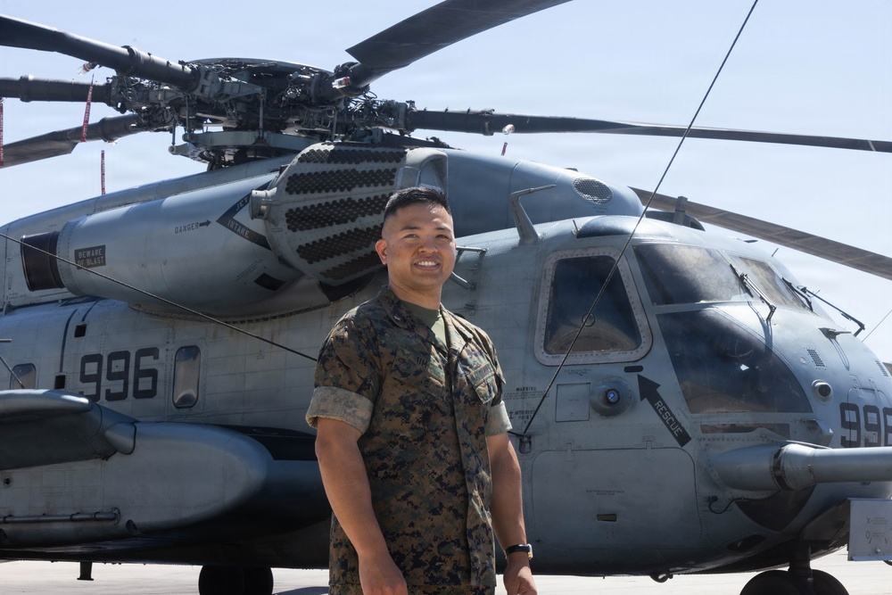 A U.S. Marine in woodland MARPAT camouflage uniform stands smiling in front of a large military helicopter on a tarmac. The helicopter bears the tail number 996 and displays markings consistent with a CH-53E Super Stallion heavy-lift helicopter. The setting appears to be a military airfield on a clear, sunny day.