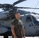 A U.S. Marine in woodland MARPAT camouflage uniform stands smiling in front of a large military helicopter on a tarmac. The helicopter bears the tail number 996 and displays markings consistent with a CH-53E Super Stallion heavy-lift helicopter. The setting appears to be a military airfield on a clear, sunny day.