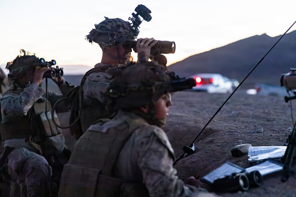 Military personnel in desert camouflage uniforms are conducting an observation or reconnaissance operation at dusk in an arid, desert environment. At least three soldiers are visible, using optical equipment including binoculars and a spotting scope to observe a distant area. A radio antenna and what appears to be a tablet or map are visible on the ground, suggesting a forward observation or fire support coordination role.