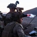 Military personnel in desert camouflage uniforms are conducting an observation or reconnaissance operation at dusk in an arid, desert environment. At least three soldiers are visible, using optical equipment including binoculars and a spotting scope to observe a distant area. A radio antenna and what appears to be a tablet or map are visible on the ground, suggesting a forward observation or fire support coordination role.