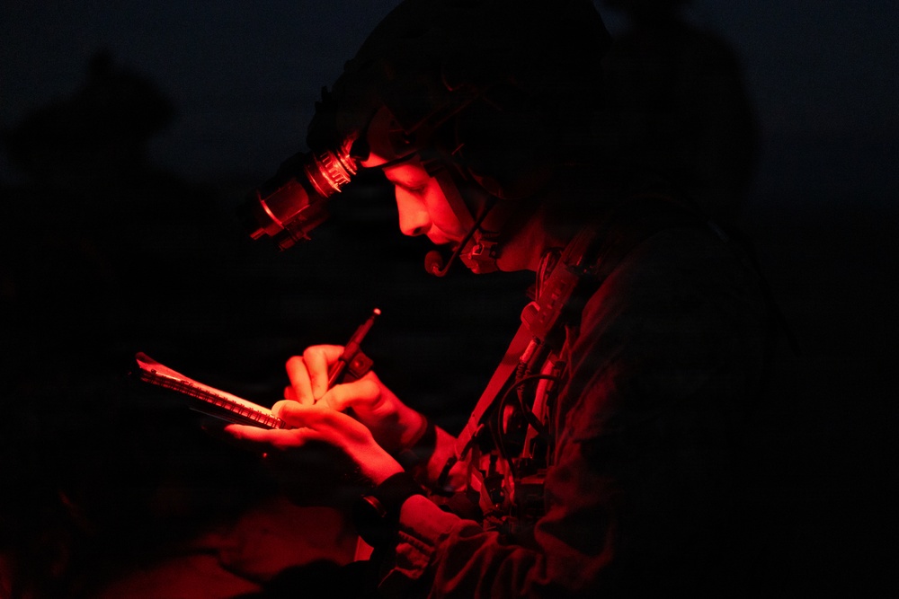 A military personnel is photographed at night under red light illumination, writing or annotating on what appears to be a tablet or notepad. The individual is wearing a tactical helmet equipped with night vision goggles (NVGs) mounted on the front. The red lighting is consistent with tactical night operations, as red light preserves night vision.