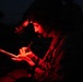 A military personnel is photographed at night under red light illumination, writing or annotating on what appears to be a tablet or notepad. The individual is wearing a tactical helmet equipped with night vision goggles (NVGs) mounted on the front. The red lighting is consistent with tactical night operations, as red light preserves night vision.