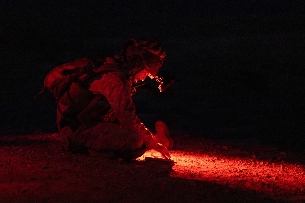 A military personnel is crouched low to the ground at night, illuminated by a red light source emanating from below. The soldier appears to be in full combat gear including helmet and tactical equipment, examining or working with an object on the ground. The scene is set in a dark, rocky outdoor environment with the red light providing the only visible illumination.