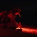 A military personnel is crouched low to the ground at night, illuminated by a red light source emanating from below. The soldier appears to be in full combat gear including helmet and tactical equipment, examining or working with an object on the ground. The scene is set in a dark, rocky outdoor environment with the red light providing the only visible illumination.