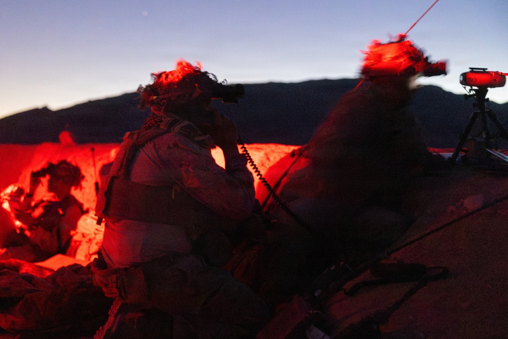 The image shows military personnel operating in low-light/dusk conditions, illuminated by a red light source. Soldiers wearing helmets with night-vision device mounts are positioned behind rocky terrain, with one holding a coiled communication cord. A tripod-mounted device is visible on the right side of the frame.