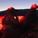 The image shows military personnel operating in low-light/dusk conditions, illuminated by a red light source. Soldiers wearing helmets with night-vision device mounts are positioned behind rocky terrain, with one holding a coiled communication cord. A tripod-mounted device is visible on the right side of the frame.