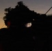 A soldier or military personnel is photographed at dusk or dawn while looking through an optical scope or sighting device. The subject wears a camouflaged helmet adorned with foliage and has hearing protection visible on the side. The dramatic low-light composition illuminates only the eyes and partial face, creating a stark contrast against the twilight sky.