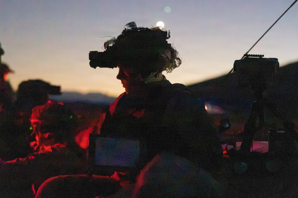 A military personnel is photographed at dusk or dawn wearing a helmet mounted with night vision goggles (NVG) and camouflage netting. The soldier appears to be at a command or observation post, surrounded by tactical equipment including a laptop and radio communications gear. The scene is dramatically lit by a twilight sky and red-tinted artificial light sources.
