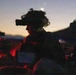 A military personnel is photographed at dusk or dawn wearing a helmet mounted with night vision goggles (NVG) and camouflage netting. The soldier appears to be at a command or observation post, surrounded by tactical equipment including a laptop and radio communications gear. The scene is dramatically lit by a twilight sky and red-tinted artificial light sources.