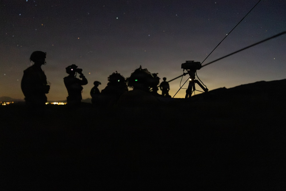 A nighttime military or surveillance operation showing multiple silhouetted personnel on elevated terrain against a twilight sky with visible stars. At least one individual is using optical equipment such as binoculars, and a tripod-mounted device with an antenna or cable is positioned on the right side. City lights are faintly visible on the horizon in the background.