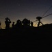 A nighttime military or surveillance operation showing multiple silhouetted personnel on elevated terrain against a twilight sky with visible stars. At least one individual is using optical equipment such as binoculars, and a tripod-mounted device with an antenna or cable is positioned on the right side. City lights are faintly visible on the horizon in the background.