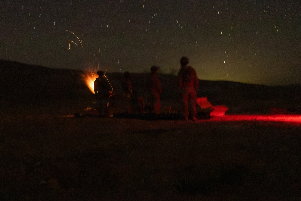 A long-exposure nighttime photograph showing several silhouetted figures gathered around what appears to be a mortar or artillery piece firing, with a visible muzzle flash or explosion of light. A red light source illuminates equipment on the right side of the frame. Star trails are visible in the sky, indicating an extended camera exposure time.