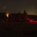 A long-exposure nighttime photograph showing several silhouetted figures gathered around what appears to be a mortar or artillery piece firing, with a visible muzzle flash or explosion of light. A red light source illuminates equipment on the right side of the frame. Star trails are visible in the sky, indicating an extended camera exposure time.