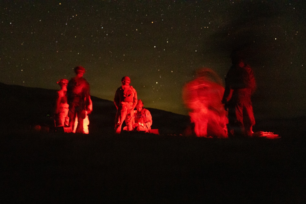 A nighttime outdoor scene showing several military personnel illuminated by red light against a starry sky. The figures appear to be engaged in ground-level activity, possibly a field operation or training exercise. The red illumination is consistent with tactical red-light usage to preserve night vision.