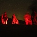 A nighttime outdoor scene showing several military personnel illuminated by red light against a starry sky. The figures appear to be engaged in ground-level activity, possibly a field operation or training exercise. The red illumination is consistent with tactical red-light usage to preserve night vision.