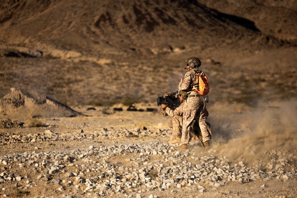 Two military personnel in desert camouflage uniforms are operating in an arid, rocky terrain. One soldier is standing and wearing a helmet with communications equipment and a bright orange backpack, while the other appears to be in a lower position handling a crew-served weapon system. Dust is visible around their feet, suggesting recent movement or weapon discharge.
