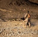 Two military personnel in desert camouflage uniforms are operating in an arid, rocky terrain. One soldier is standing and wearing a helmet with communications equipment and a bright orange backpack, while the other appears to be in a lower position handling a crew-served weapon system. Dust is visible around their feet, suggesting recent movement or weapon discharge.