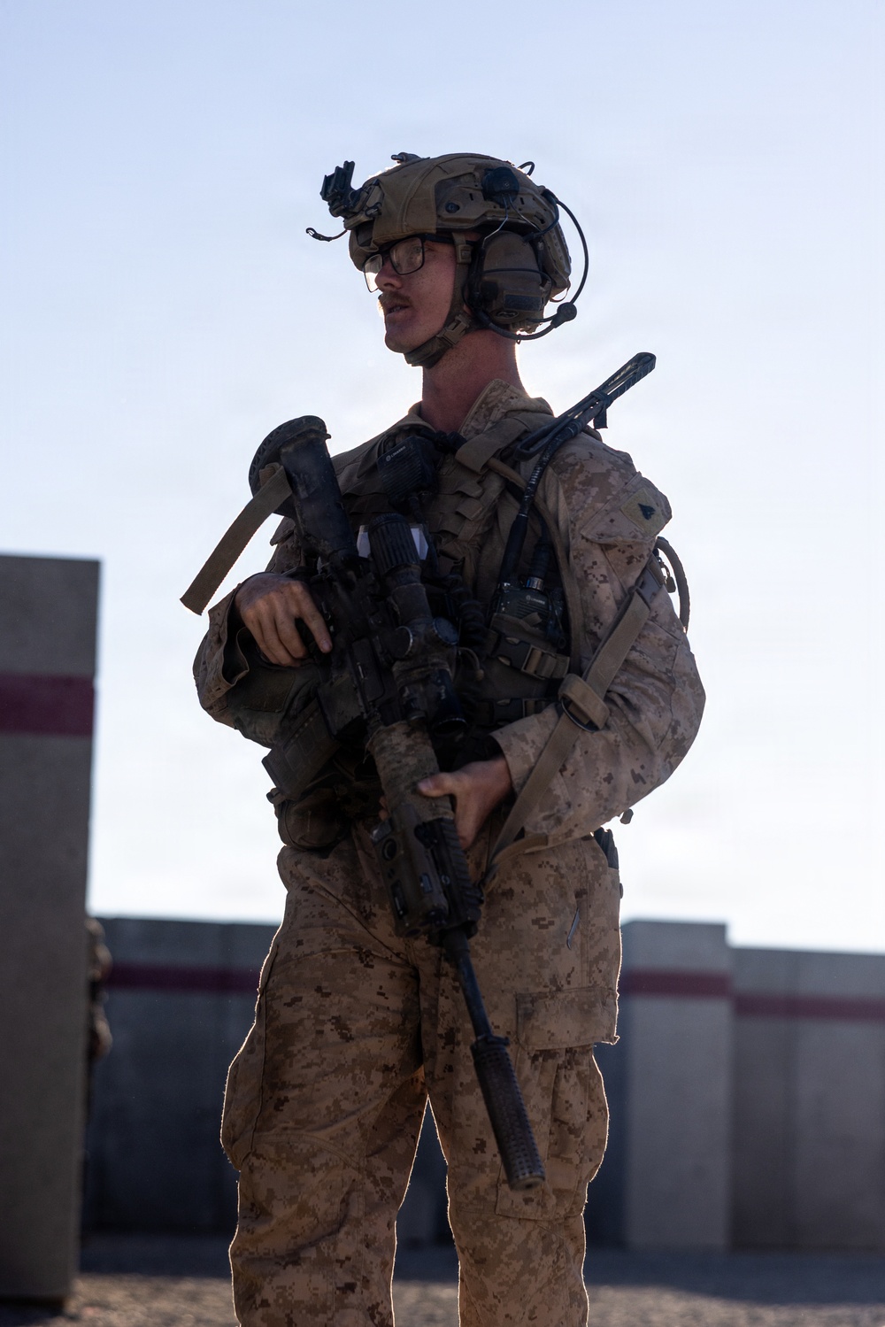 A U.S. Marine in full combat gear stands in an outdoor environment, holding an assault rifle in a ready position. The soldier is equipped with advanced tactical equipment including a helmet-mounted night vision device, communications headset, and body armor. The setting appears to be a training or operational area with concrete barriers visible in the background.