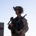 A U.S. Marine in full combat gear stands in an outdoor environment, holding an assault rifle in a ready position. The soldier is equipped with advanced tactical equipment including a helmet-mounted night vision device, communications headset, and body armor. The setting appears to be a training or operational area with concrete barriers visible in the background.