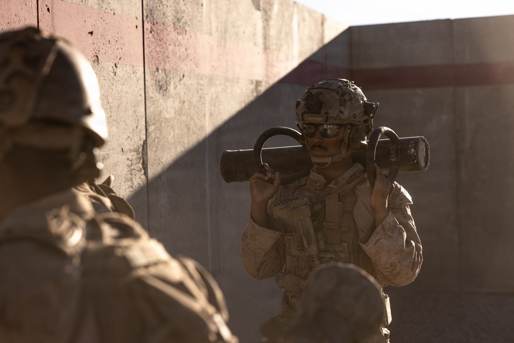The image shows military personnel in desert camouflage uniforms operating in what appears to be an urban or compound environment with concrete walls. The central figure is carrying a shoulder-launched rocket/anti-tank weapon system on their shoulder. The scene suggests a training exercise or operational deployment in an arid environment.