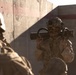 The image shows military personnel in desert camouflage uniforms operating in what appears to be an urban or compound environment with concrete walls. The central figure is carrying a shoulder-launched rocket/anti-tank weapon system on their shoulder. The scene suggests a training exercise or operational deployment in an arid environment.
