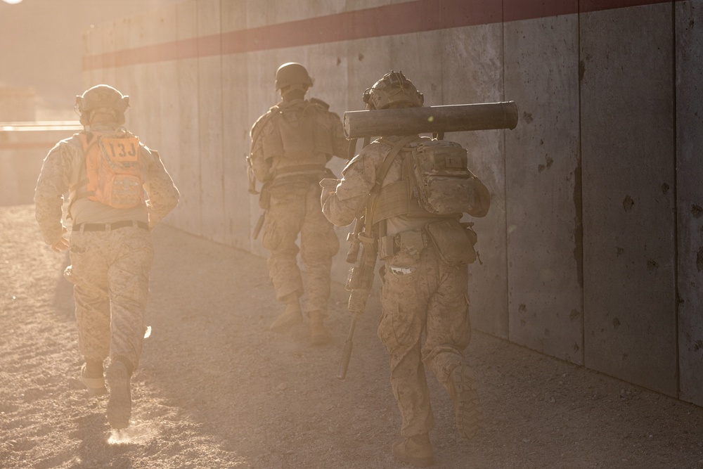 Three military personnel in desert camouflage uniforms are moving along a concrete barrier wall in dusty, hazy conditions. One soldier on the left wears an orange vest marked '13J', suggesting a training or exercise role identifier. The soldier on the right carries what appears to be a shoulder-launched weapon system (likely an AT4 or similar rocket launcher).