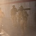 Three military personnel in desert camouflage uniforms are moving along a concrete barrier wall in dusty, hazy conditions. One soldier on the left wears an orange vest marked '13J', suggesting a training or exercise role identifier. The soldier on the right carries what appears to be a shoulder-launched weapon system (likely an AT4 or similar rocket launcher).