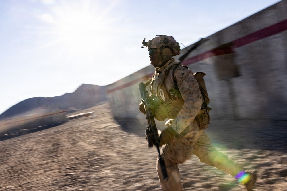 A fully equipped soldier in desert camouflage (MARPAT pattern) is running at speed past a concrete wall with a red stripe, in an arid, desert environment. The motion blur effect conveys rapid movement and urgency. The setting appears to be a training facility or operational area with mountainous terrain visible in the background.
