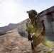 A fully equipped soldier in desert camouflage (MARPAT pattern) is running at speed past a concrete wall with a red stripe, in an arid, desert environment. The motion blur effect conveys rapid movement and urgency. The setting appears to be a training facility or operational area with mountainous terrain visible in the background.