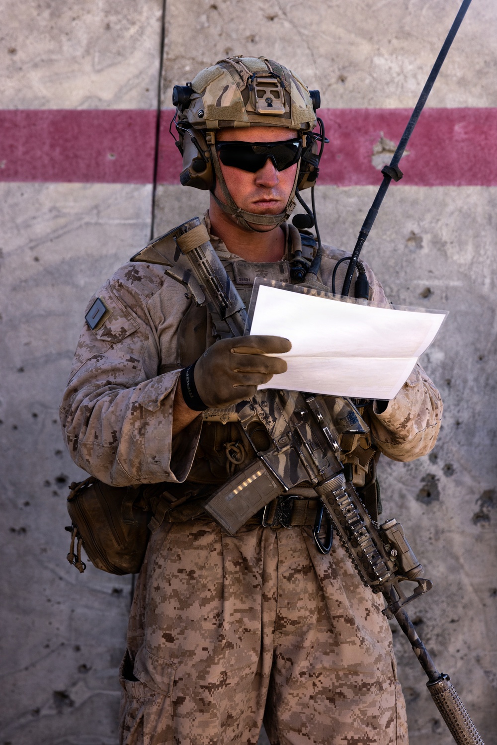 A U.S. Marine in full combat gear stands against a concrete wall with a red stripe, reading a document. The Marine is equipped with MARPAT desert camouflage, a tactical helmet with communications equipment, and an M4-style carbine. The setting appears to be an operational environment, likely in a Middle Eastern or arid region.