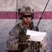 A U.S. Marine in full combat gear stands against a concrete wall with a red stripe, reading a document. The Marine is equipped with MARPAT desert camouflage, a tactical helmet with communications equipment, and an M4-style carbine. The setting appears to be an operational environment, likely in a Middle Eastern or arid region.