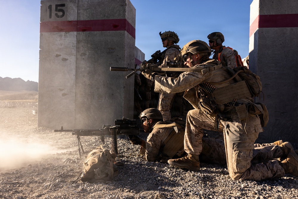 A group of military personnel in desert camouflage uniforms are conducting what appears to be a tactical training exercise at a range facility. Soldiers are positioned around concrete barrier structures marked with red stripes and the number '15', with at least one machine gun actively firing. The arid, mountainous background and controlled range environment strongly suggest a military training installation.