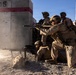 A group of military personnel in desert camouflage uniforms are conducting what appears to be a tactical training exercise at a range facility. Soldiers are positioned around concrete barrier structures marked with red stripes and the number '15', with at least one machine gun actively firing. The arid, mountainous background and controlled range environment strongly suggest a military training installation.
