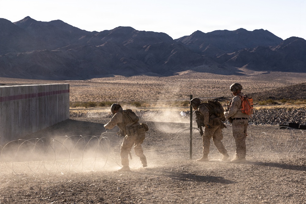 Three military personnel in desert camouflage uniforms are conducting a training exercise in an arid desert environment. They appear to be breaching or navigating through concertina/razor wire obstacles near a concrete wall structure. One individual wearing an orange safety vest is likely an observer or safety officer overseeing the exercise.