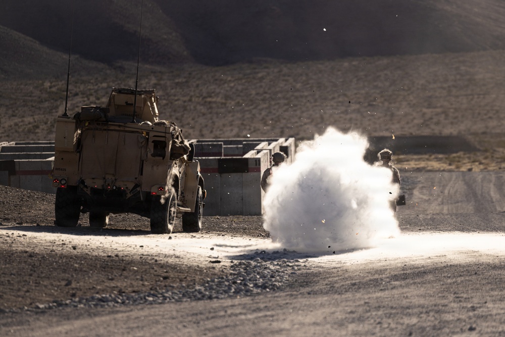 A military training or operational scenario in an arid, desert environment showing a large explosion or detonation on a gravel road. A tan/sand-colored military tactical vehicle (JLTV or similar MRAP-type vehicle) is positioned to the left, while armed personnel in combat gear take cover behind concrete barriers to the right. The scene appears to be a controlled demolition exercise or simulated IED scenario based on the structured environment.