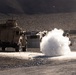 A military training or operational scenario in an arid, desert environment showing a large explosion or detonation on a gravel road. A tan/sand-colored military tactical vehicle (JLTV or similar MRAP-type vehicle) is positioned to the left, while armed personnel in combat gear take cover behind concrete barriers to the right. The scene appears to be a controlled demolition exercise or simulated IED scenario based on the structured environment.
