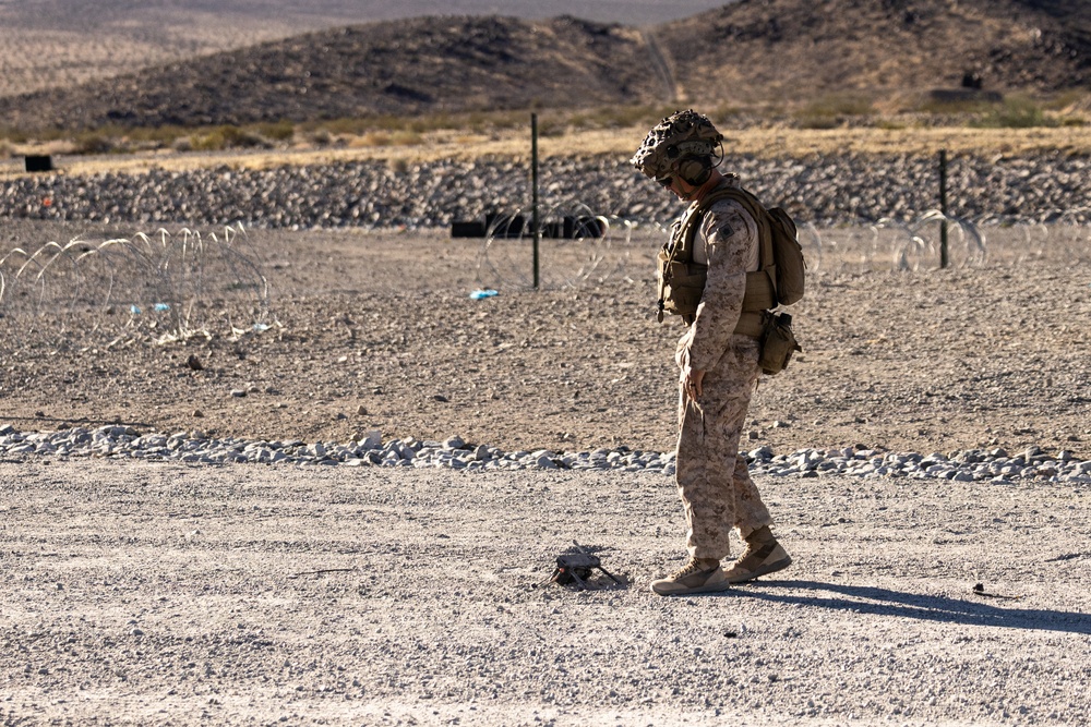 A U.S. Marine in full combat gear, wearing desert MARPAT camouflage, stands in an arid desert training environment looking down at an object on the ground. The setting appears to be a military training range, evidenced by concertina wire barriers, rock berms, and range markers in the background. The terrain and vegetation are consistent with a Mojave Desert location, likely a base such as Twentynine Palms (MCAGCC).