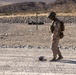 A U.S. Marine in full combat gear, wearing desert MARPAT camouflage, stands in an arid desert training environment looking down at an object on the ground. The setting appears to be a military training range, evidenced by concertina wire barriers, rock berms, and range markers in the background. The terrain and vegetation are consistent with a Mojave Desert location, likely a base such as Twentynine Palms (MCAGCC).