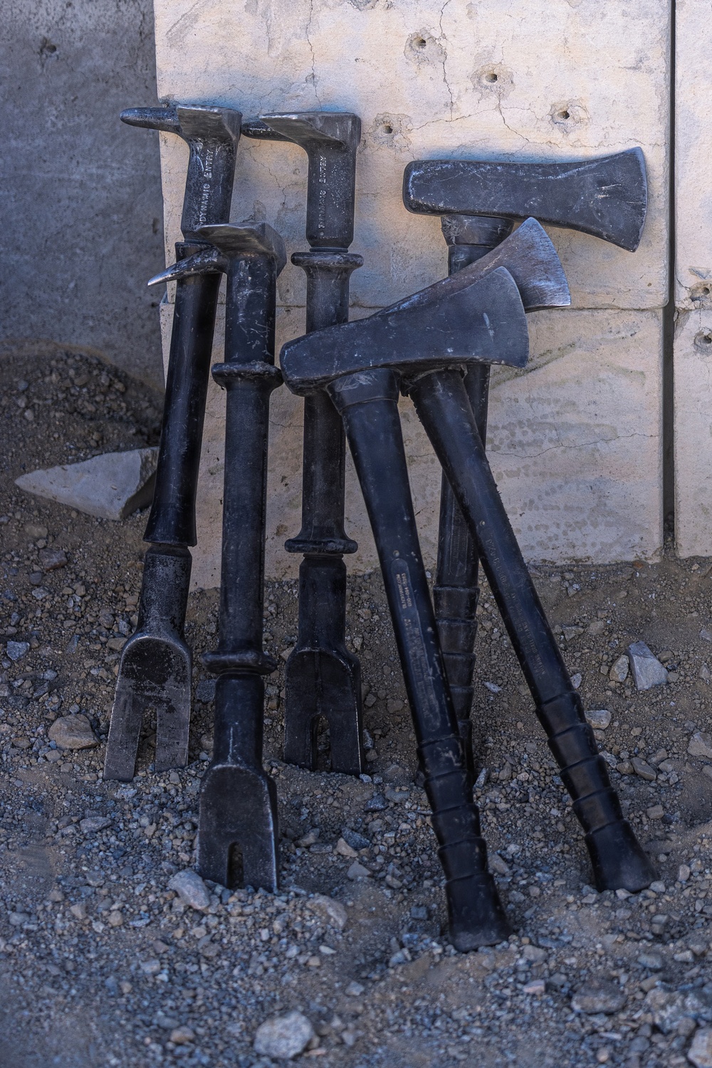 A collection of heavy breaching and entry tools leaned against a concrete wall on a gravel/dirt surface. The tools include Halligan bars, axes, and sledgehammer-type implements, all finished in matte black. Text visible on at least two of the tools reads 'DYNAMIC ENTRY', indicating these are tactical law enforcement or military breaching tools.