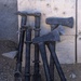 A collection of heavy breaching and entry tools leaned against a concrete wall on a gravel/dirt surface. The tools include Halligan bars, axes, and sledgehammer-type implements, all finished in matte black. Text visible on at least two of the tools reads 'DYNAMIC ENTRY', indicating these are tactical law enforcement or military breaching tools.