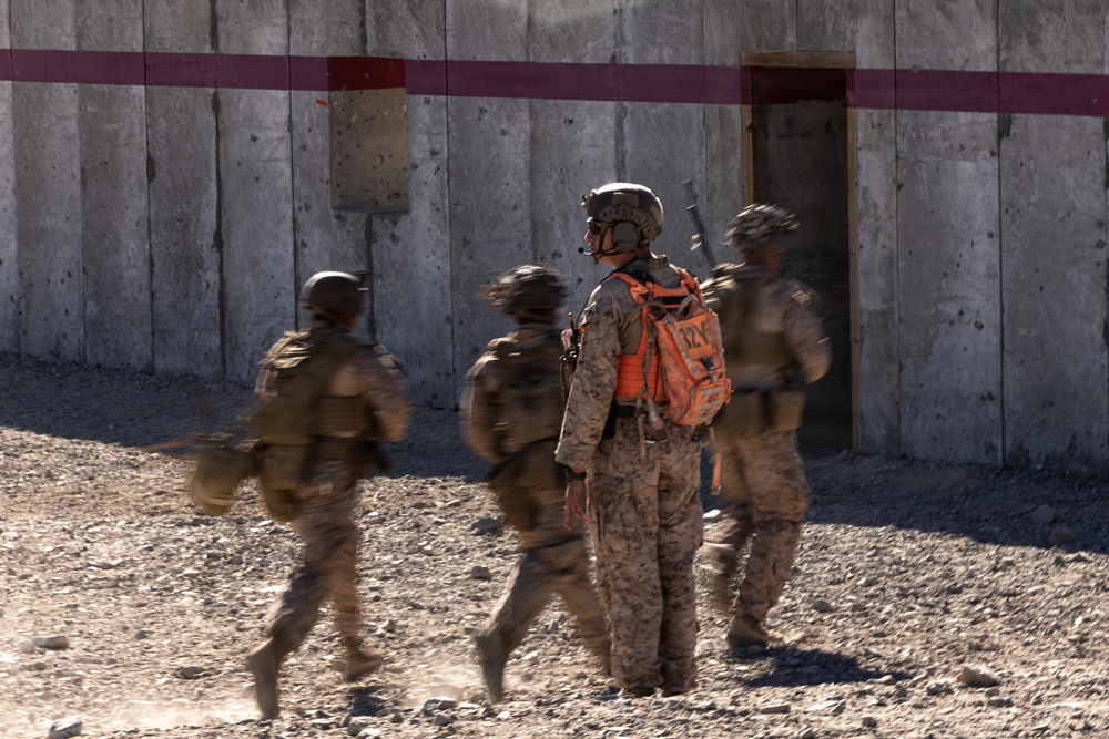 The image shows a group of military personnel in desert camouflage uniforms approaching a concrete structure with a doorway. One central figure wearing an orange safety/observer vest and helmet stands stationary while others move around them, creating motion blur. The setting appears to be a military training exercise based on the orange vest worn by the observer/safety officer.