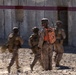 The image shows a group of military personnel in desert camouflage uniforms approaching a concrete structure with a doorway. One central figure wearing an orange safety/observer vest and helmet stands stationary while others move around them, creating motion blur. The setting appears to be a military training exercise based on the orange vest worn by the observer/safety officer.