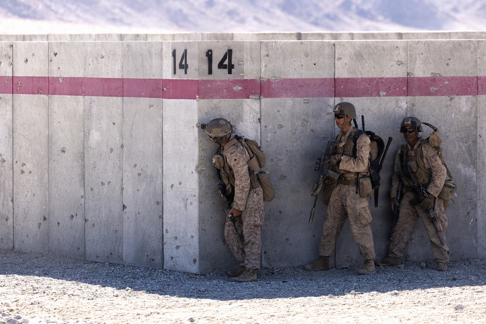 Three military personnel in desert camouflage uniforms are positioned against a concrete barrier wall marked with the number '14' and a horizontal red/pink stripe. One soldier is leaning around the corner of the wall in a tactical peek position, while the other two stand against the wall holding rifles. The setting appears to be an arid, desert training or operational environment with mountains visible in the background.