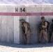 Three military personnel in desert camouflage uniforms are positioned against a concrete barrier wall marked with the number '14' and a horizontal red/pink stripe. One soldier is leaning around the corner of the wall in a tactical peek position, while the other two stand against the wall holding rifles. The setting appears to be an arid, desert training or operational environment with mountains visible in the background.