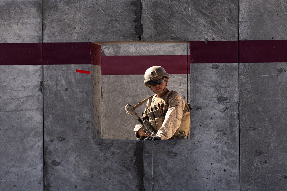 A soldier in full combat gear is visible through a square opening in a large concrete wall marked with dark red/maroon painted stripes. The soldier is wearing a tan/desert camouflage uniform, tactical vest, helmet, and sunglasses while holding a rifle. The setting appears to be a military installation or training facility.
