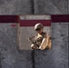 A soldier in full combat gear is visible through a square opening in a large concrete wall marked with dark red/maroon painted stripes. The soldier is wearing a tan/desert camouflage uniform, tactical vest, helmet, and sunglasses while holding a rifle. The setting appears to be a military installation or training facility.