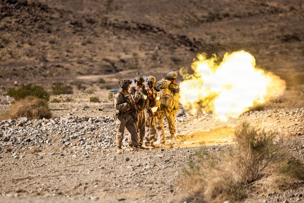 A group of military personnel in desert camouflage uniforms are operating what appears to be a mortar or similar crew-served weapon in an arid, rocky desert environment. A large fireball and muzzle blast are visible immediately to the right of the group, indicating a weapon has just been fired. The terrain and vegetation suggest a desert training range environment.