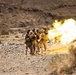 A group of military personnel in desert camouflage uniforms are operating what appears to be a mortar or similar crew-served weapon in an arid, rocky desert environment. A large fireball and muzzle blast are visible immediately to the right of the group, indicating a weapon has just been fired. The terrain and vegetation suggest a desert training range environment.