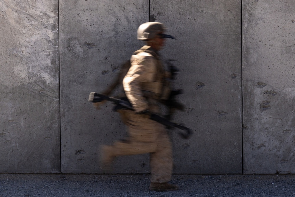 A motion-blurred image of a soldier in full combat gear running past a large concrete wall. The deliberate slow-shutter photography technique conveys speed and urgency. The subject is moving from left to right across the frame.