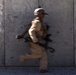 A motion-blurred image of a soldier in full combat gear running past a large concrete wall. The deliberate slow-shutter photography technique conveys speed and urgency. The subject is moving from left to right across the frame.